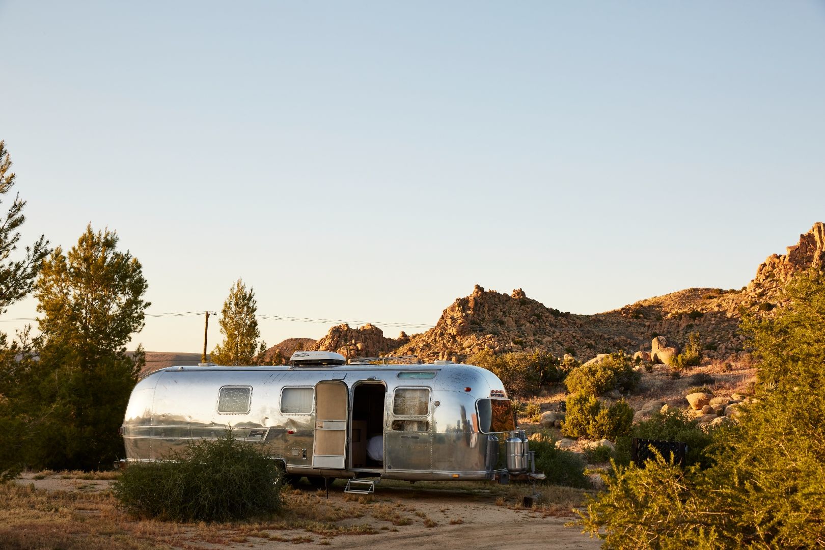 Large Airstream at ESCAPE — side-on at golden hour, framed by junipers and the boulder ridge