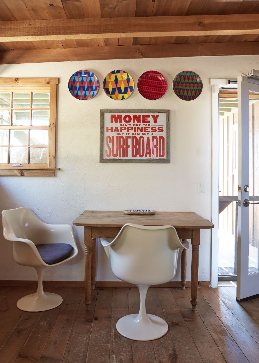 The Residence dining nook at ESCAPE — tulip table and chairs under a pine-beam ceiling, decorative wall plates and the &lsquo;Money can&rsquo;t buy you happiness but it can buy a Surfboard&rsquo; letterpress poster