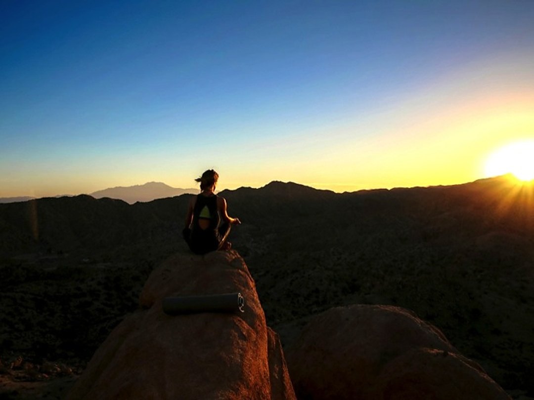 A woman meditating on a boulder above ESCAPE at sunset, a yoga mat beside her